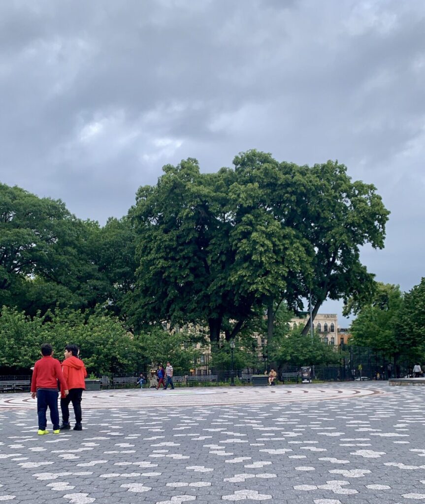 A picture from Maria Hernandez Park's center circle on an overcast day. Two boys in red hoodies are in the foreground with their backs to the camera, while other neighbors can be seen sitting on benches or walking through the park in the background.